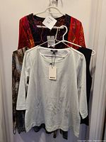 Four women's tops hanging on white plastic and metal hangers, size medium, different colors and patterns visible, some with tags attached.