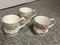 Three bone china teacups with floral designs placed on grey carpet showing shape and handle detail.