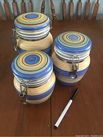 Three ceramic canisters with clip-top lids and colorful horizontal stripes in blue, green, yellow, orange, and red shades placed on a wooden table, alongside a marker for scale.