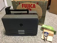 Side view of the Fujica 8 projector, its original box behind it, and the spare lamp bulb in packaging beside it on a carpeted floor.