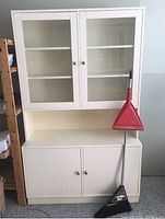 Full view of the closed two-piece laminate hutch with glass upper doors and solid lower doors. A red dustpan and brush set is leaning against the right side.