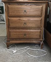 Front view photo of a solid wood side table with three drawers and metal ring pulls on carpeted floor, electrical cords in front.