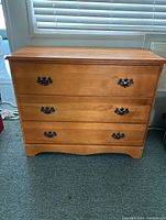 Front view of a pine dresser with three drawers and brass handles, placed on a carpeted floor in front of a window with blinds.