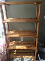Front view of a five-tier pine wood shelving unit illuminated by natural light. Shelves are open with slatted surfaces and some wood knots visible.