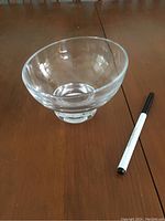 Clear glass bowl with angled rim and pedestal base shown on wooden table with pen for scale.