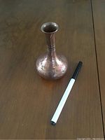 Photo of a small bulbous copper-colored metal vase with narrow neck and flared top, placed on wooden surface with a marker pen for scale.
