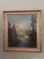 Framed vintage oil painting showing a landscape scene with trees, water, mountains, and a house. Frame is gold with cream border.