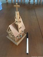 Photo of a small light brown wooden church model on a wooden table. The model has a tall steeple with a golden cross on top, arched windows, and a detailed roof structure. A pen is placed next to it for scale.