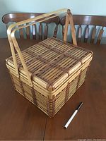 Woven picnic basket closed on a wooden table with pen for size reference
