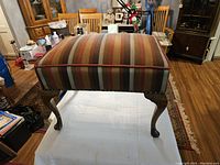 Front view of an antique ottoman foot stool showing rectangular shape, striped fabric upholstery with multiple colors, and wooden cabriole legs.
