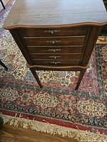 Full view of the jewelry armoire showing its wooden body, curved top, metal handles on five visible drawers, and standing legs on a patterned rug.