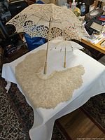Adult-sized Battenberg lace parasol, doll-sized lace parasol, and Victorian lace bed jacket displayed on a table with white cloth.