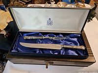 Full view of carving set knife and fork inside open presentation box with purple satin lining and Birks logo.