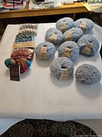 Overview photo showing 9 large balls of light blue/grey Drops Air mix yarn and 10 mixed skeins of Manos del Uruguay Fino fingering wool yarn in multiple colors.