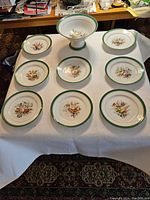 Overview of all 8 dessert plates and a footed fruit bowl laid out on a white cloth