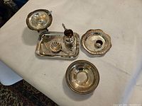 Overview of nine silver plated serving pieces arranged on a beige fabric surface including trays, bowls, and a small pitcher.