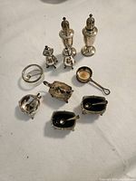 Wide shot showing entire set of silver tableware including salt cellars, pepper shakers, ladle, and glass piece on white cloth surface.