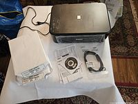 Canon PIXMA MG3600 series printer on white table with two packages of 8.5x11 copy paper wrapped in plastic, power cord, instruction sheets, driver CD and USB cable.