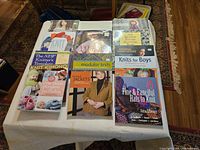 Photo showing 13 colorful knitting books arranged on a table, covers showing knitting projects including hats, jackets, kimonos, mitts, and boys' wear.