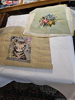 Two finished needlepoint pieces laid flat: one with a floral bouquet on pale green background, the other with a cat portrait on beige background.