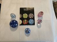 Five distinct glass paperweights arranged around a book on a white cloth surface showing the overall lot contents.