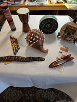 Full overview of assorted percussion instruments laid on white tablecloth showing djembe, flutes, shekere, wooden spoons, slit drum and steel tongue drum