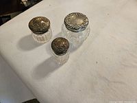 Three small vintage clear glass cosmetic vessels with detailed sterling silver lids displayed on a white cloth surface.