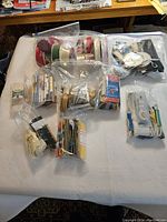 Full lot overview showing multiple plastic bags containing sewing supplies such as ribbons and threads, assorted needles, and pins on a white surface.