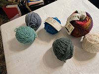 Six balls of worsted wool yarn in assorted colors placed on a white cloth: cream, light teal, medium gray, dark gray, blue, and multicolor maroon-red.