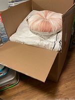 Open cardboard box showing folded white bedding items and a peach decorative round cushion on top