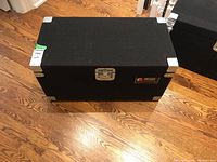 Closed Odyssey storage trunk with black exterior, metal hardware, and coat of arms logo plate on front.