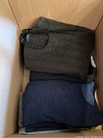 Three folded sweaters in box: olive cable knit, charcoal crewneck, heather gray