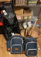 Two Happy Picnic brand navy blue picnic backpacks with tan trim, shopper cart with black quilted synthetic bag and metal frame with wheels, three wicker baskets in front