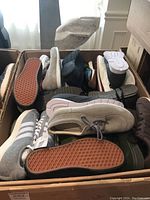 Top view of a box containing multiple pairs of shoes in various colors and styles including grey, black, and white casual and athletic footwear.