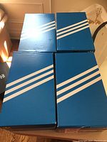 Four blue Adidas shoeboxes stacked on floor showing the lot includes 4 pairs of Adidas shoes.