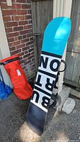 Snowboard leaning against fence with red bag in background, showing overall board graphic and bag.