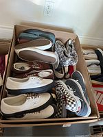 Top view photo of a box of approximately 15 pairs of Vans shoes with multiple different designs and colors visible, including black and white checkers and solid white with black soles.