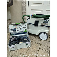 Festool shop vacuum with green hose and wheeled base alongside open Systainer case containing Festool Rotex sander and accessories on paved surface.