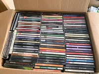 Photo showing one of four boxes open with rows of jewel case CDs packed inside, titles visible including various artists and albums in different musical genres.