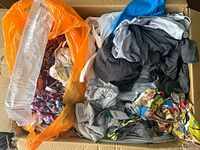 Top view of a cardboard box filled with assorted clothing for women, men, and children, showing two orange bags – one with a visible receipt and clothing including grey and patterned items