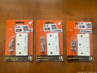 Photo shows three unopened packages of Hillman 3 inch (76 mm) white door bracket hinges, each with screws included, priced $5.48 each.