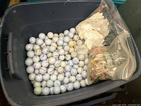 Large black plastic bin filled with numerous white golf balls with various logos, several golf gloves on top, and bags with colorful golf tees.