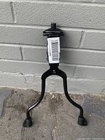 Front view of black metal bicycle kickstand against gray brick wall, showing two legs with rubber feet and top mounting plate.