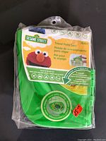 Photo of green foldable Sesame Street travel potty seat in sealed plastic packaging. Packaging shows Elmo and indicates suitability for children 18+ months.