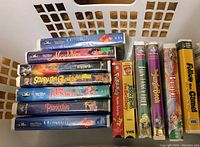 Image shows a stack of twelve kids VHS tapes in a laundry basket. Titles include mostly Disney movies such as The Little Mermaid, Aladdin, Pinocchio, The Rescuers, The Jungle Book, Bambi, plus Scooby-Doo! Ghoul School, Pokémon, and others.
