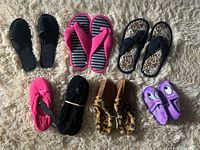 Eight pairs of new women's and kids' slippers and sandals arranged on a beige rug including black slip-ons, pink flip-flops, leopard-print sandals, bright pink fleece slippers, black strap slippers, brown fleece slippers, and purple ones with eyes.