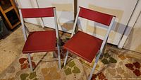 Two red folding chairs with plastic seats and backs on metal frames positioned on a floral carpet.
