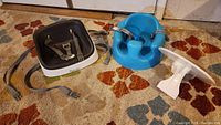 Photo showing blue Bumbo infant floor seat with attached white plastic tray, and black and white Ingenuity booster seat with gray removable pad and safety straps on carpeted floor.