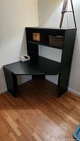 Black corner desk with triangular surface and one-sided hutch shelf with two cubbies on top, positioned in room corner over hardwood floor.