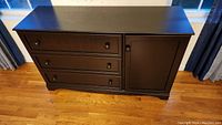 Front view of black laminated particle board dresser with three drawers and one door with knobs